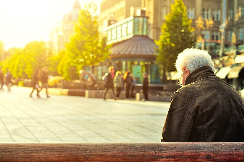 Senior with white hair sitting alone on a bench in a sunny public square, with people and buildings in the blurred background
